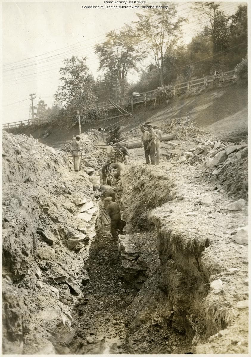 Workers standing in a deep, narrow excavation trench cut through open ground, with earth piled along its edges.