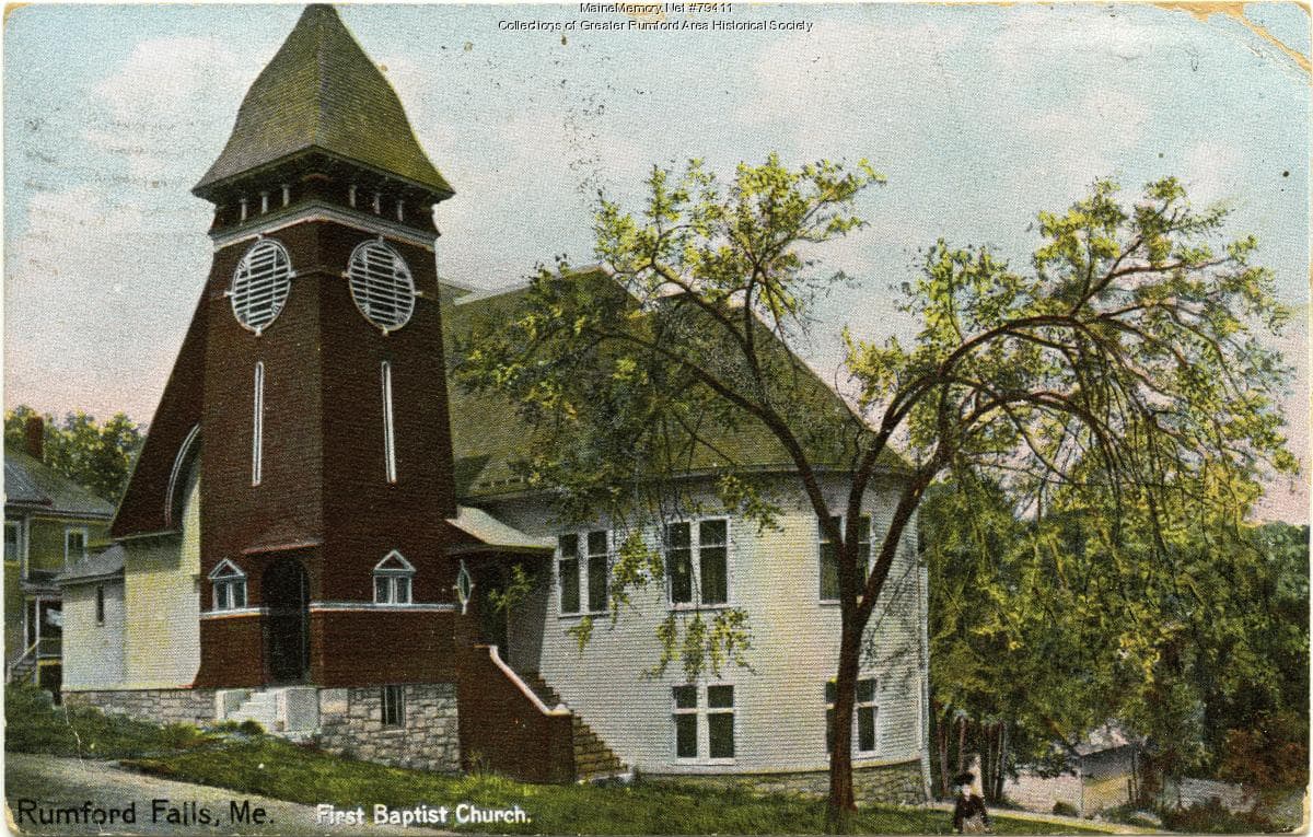 A church building with a corner bell tower, photographed for a postcard from across the street.