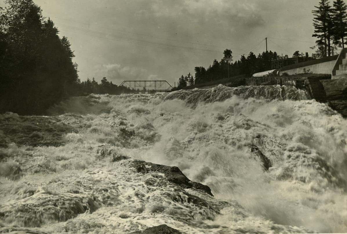 The Androscoggin River in full flood over Rumford Falls, with a steel truss bridge on the horizon and mill retaining walls along the far bank.