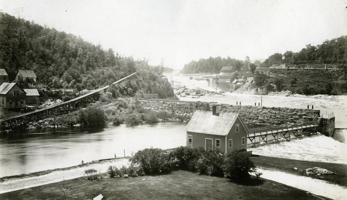 A wooden log sluice running down a wooded hillside to the river, with mill buildings and a dam beyond.