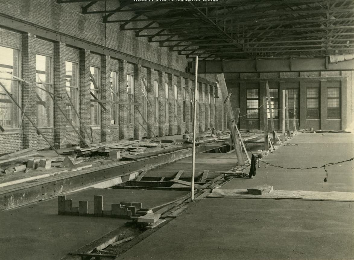 The interior of a long brick mill hall under construction, with steel roof trusses overhead and timber shoring along a wall of windows.
