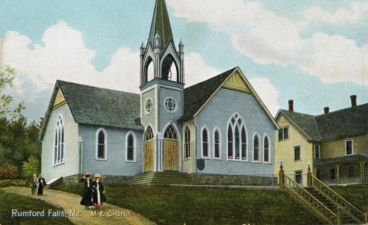 A hand-coloured postcard of a pale blue clapboard church with a tall steeple, women in long dresses walking on the path outside.