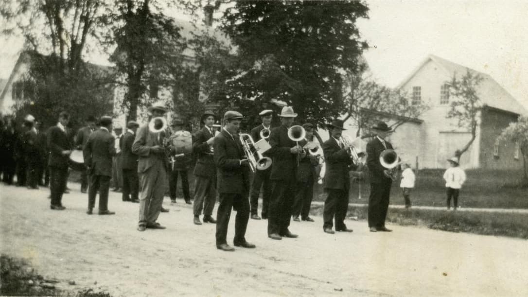 A brass band in suits and hats standing in a dirt road during a parade, with onlookers and farm buildings behind them.