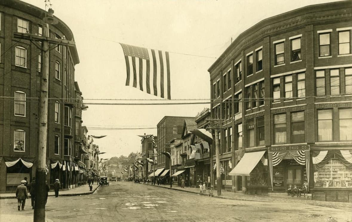 A downtown street corner lined with brick commercial blocks, an American flag hung across the street, and pedestrians on the sidewalks.